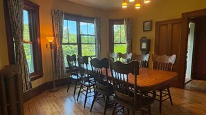 A dining area with a wooden table and chairs, large windows with curtains, and a wall-mounted clock. - The Owl's Roost Manor in Owl's Head, ME (Owls Head)