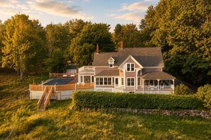 A two-story house with a porch, a fenced deck, and a stone wall. - The Owl's Roost Manor in Owl's Head, ME (Owls Head)