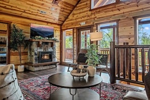 Ein Holzblockhaus-Interieur mit Kamin aus Stein, einem Fernseher und einem Balkon mit Geländer.