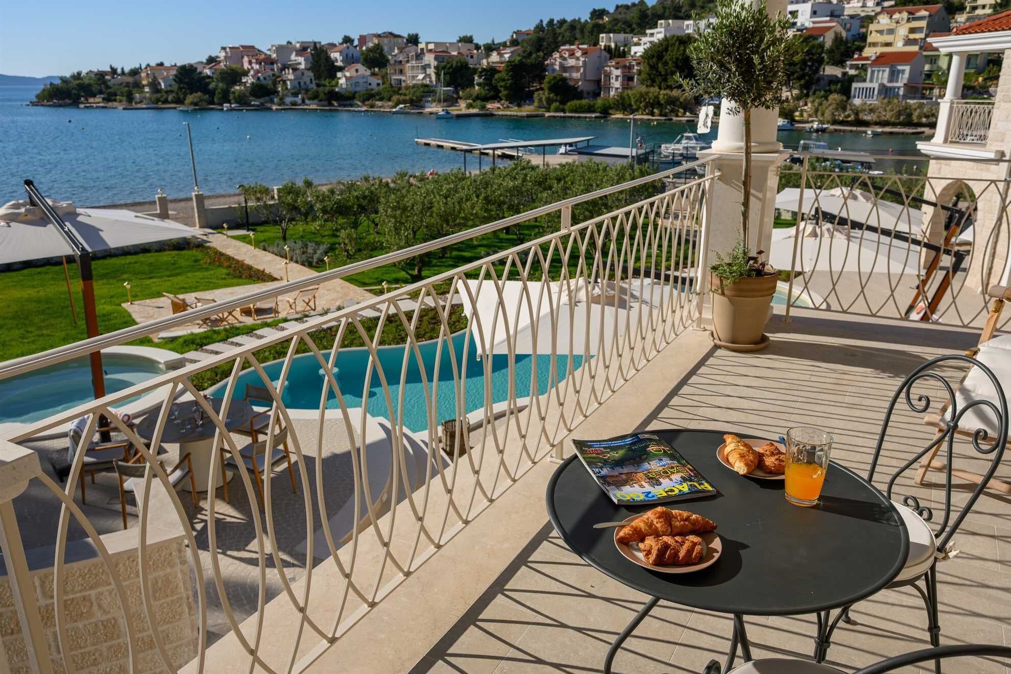 A balcony with a pool, dining table, and chairs, overlooking a coastal town.