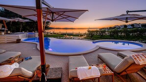 Uma área na piscina com espreguiçadeiras, guarda-sóis e vista para o pôr do sol sobre a água.