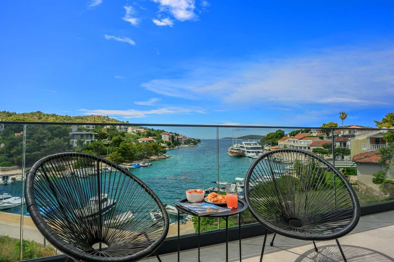 A balcony with two black wire chairs, a small table with a drink and snacks, and a view of a river with boats and buildings.