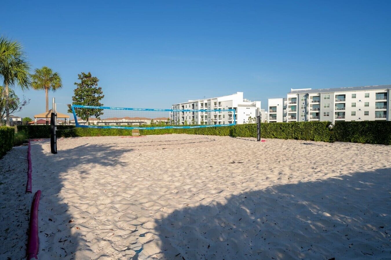 A sandy beach volleyball court with a net, surrounded by buildings and palm trees.