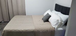Ein ordentlich bezogenes Bett mit einer beigen Tagesdecke und Kissen, ein dunkles Kopfteil und ein zur Seite gezogen Vorhang.