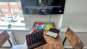 A room with a TV displaying a software upgrade message, a wooden table with board games, and a window with a view of a street. - London Southeast Modern Stay (London)