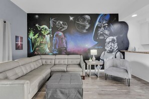 Ein modernes Wohnzimmer mit einer Eckcouch, einem grauen Hocker, einem kleinen Beistelltisch mit einer Lampe und einer Wandmalerei mit Star Wars Figuren und Raumschiffen.