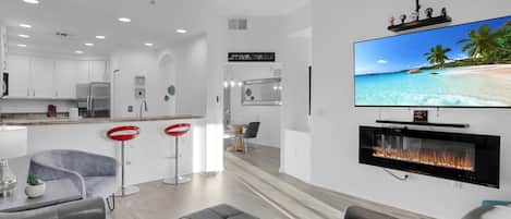 A modern living room with a flat-screen TV, a fireplace, and a kitchen area with white cabinets and red bar stools.