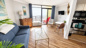 A modern living room with a sofa, a glass coffee table, and a dining area with chairs. There is a kitchen with a stove and oven. - Captivating Oceanview Balcony & Resort Pools (Myrtle Beach)