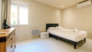 A bedroom with a bed, a dresser, a window with curtains, an air conditioner, and a small bedside table. - Opal 11 Apartment (Cannes)