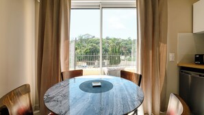 A room with a round table, chairs, a view through a window with curtains, and a kitchen area. - Opal 11 Apartment (Cannes)