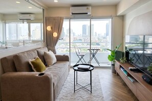 A modern living room with a sofa, a small table, a TV, and a balcony with a city view. - 49sqm 1BR in Thonglor, pool and workspace (Watthana)