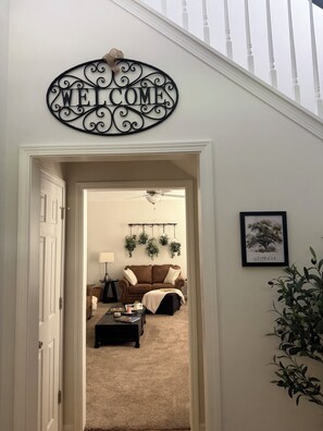 A hallway with a 'WELCOME' sign, a brown sofa, a ceiling fan, and a framed picture on the wall.
