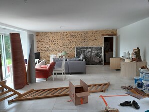 A living room with a sofa, chairs, a coffee table, a stone wall, and a cityscape painting. - Ancienne longère de caractère rénovée à 40mn du Mans (Saint-Denis-d'Orques)
