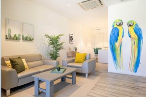 A modern living room with a sofa, armchairs, a coffee table, and a large wall painting of parrots. - Ocean View 2BR, Palm Beach Strip (Noord)