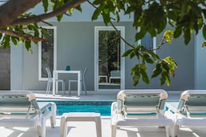 Un espace piscine avec des chaises longues, une table et des chaises, ainsi qu’un bâtiment doté de grandes fenêtres.
