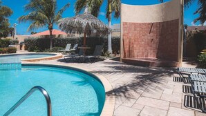 Una piscina con sillas de descanso, zona sombreada y palmeras.