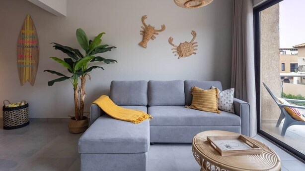 Una sala de estar moderna con un sofá gris, una planta grande en maceta, una mesa de centro de mimbre y una tabla de surf colgada en la pared.