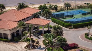A building with a red-tiled roof, surrounded by palm trees and a tennis court. - Gated Gold Coast 1BR, 75 m², 3 pools, near Malmok (Noord)