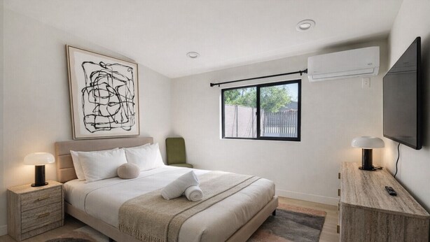 Un dormitorio con cama, mesitas de noche, televisión, ventana y un cuadro.