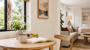 Una sala de estar acogedora con una mesa redonda de madera, un sillón cómodo, un cuenco de frutas, un libro y un jarrón con flores.