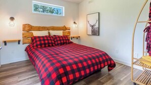 A bedroom with a wooden headboard, a checkered bedspread, a window with a view, and a wall-mounted shelf with a lamp. - Le Clorane | spa | relax (Matawinie Regional County Municipality)