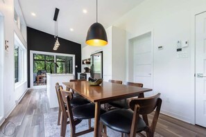 Ein moderner Essbereich mit einem Holztisch und Stühlen, einer Pendelleuchte und Blick in die Küche durch eine Glastür.
