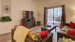 Ein Wohnzimmer mit Deckenventilator, Flachbildfernseher, Rattanmöbel, einem Glastisch und einer Schiebetür zu einer Außenpatio.