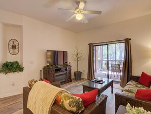 Ein Wohnzimmer mit Deckenventilator, Flachbildfernseher, Rattanmöbel, einem Glastisch und einer Schiebetür zu einer Außenpatio.