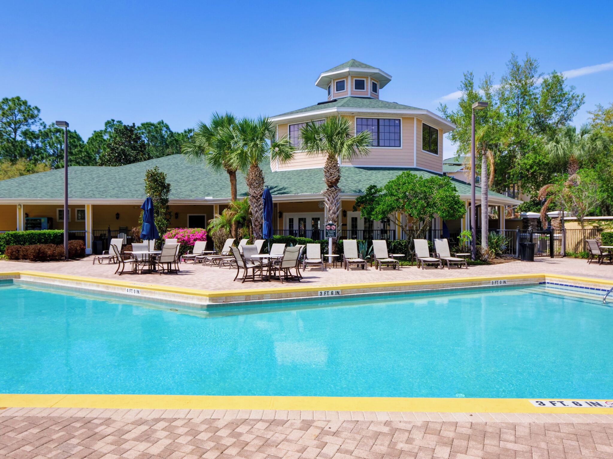 A hotel with a swimming pool, lounge chairs, palm trees, and a covered patio area.
