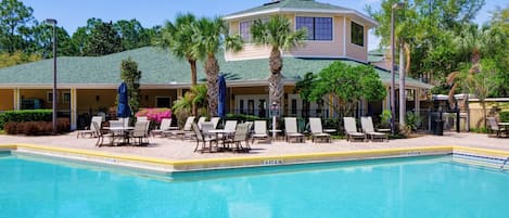A hotel with a swimming pool, lounge chairs, palm trees, and a covered patio area.