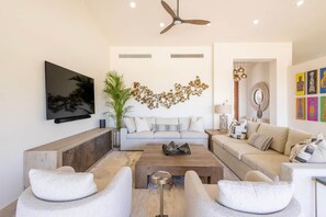 A modern living room with a sofa, coffee table, and chairs, a flat-screen TV, decorative wall art, and a ceiling fan. - Luxury Mediterranean Villa in Querencia (San José del Cabo)
