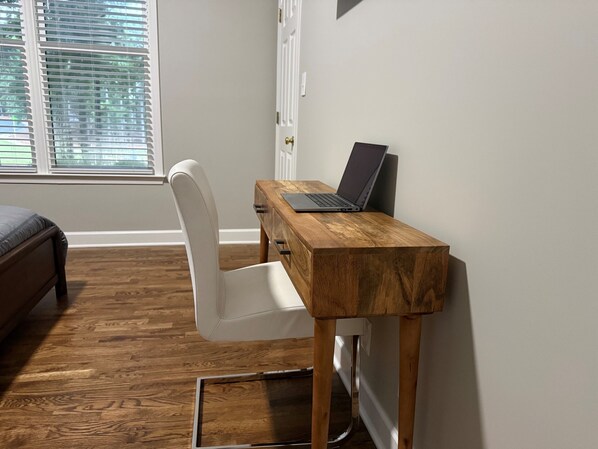 A room with a wooden desk, a laptop, a white chair, and a bed.