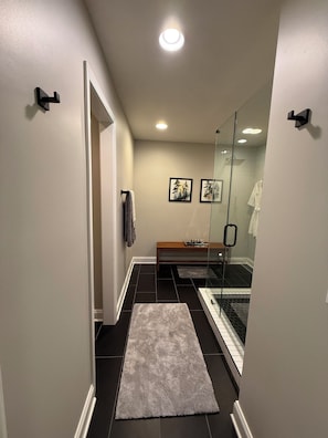 A narrow hallway with a glass shower enclosure, a wooden bench, and a gray rug on the floor.