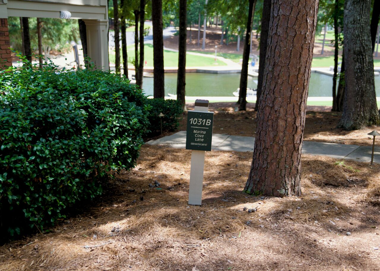 A signpost with the text '1031B Marina Cove Lane (downstairs)' in a landscaped area with trees and a grassy background.