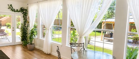 Ein Zimmer mit Glastisch, vier Stühlen, weißen Vorhängen, einem Blumentopf und Blick auf einen Garten.