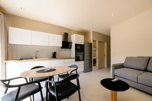 Eine moderne Küche mit weißen Schränken, einem eingebauten Ofen und einem Kühlschrank. Es gibt einen Essbereich mit einem runden Tisch und Stühlen, sowie ein graues Sofa im Wohnzimmer.