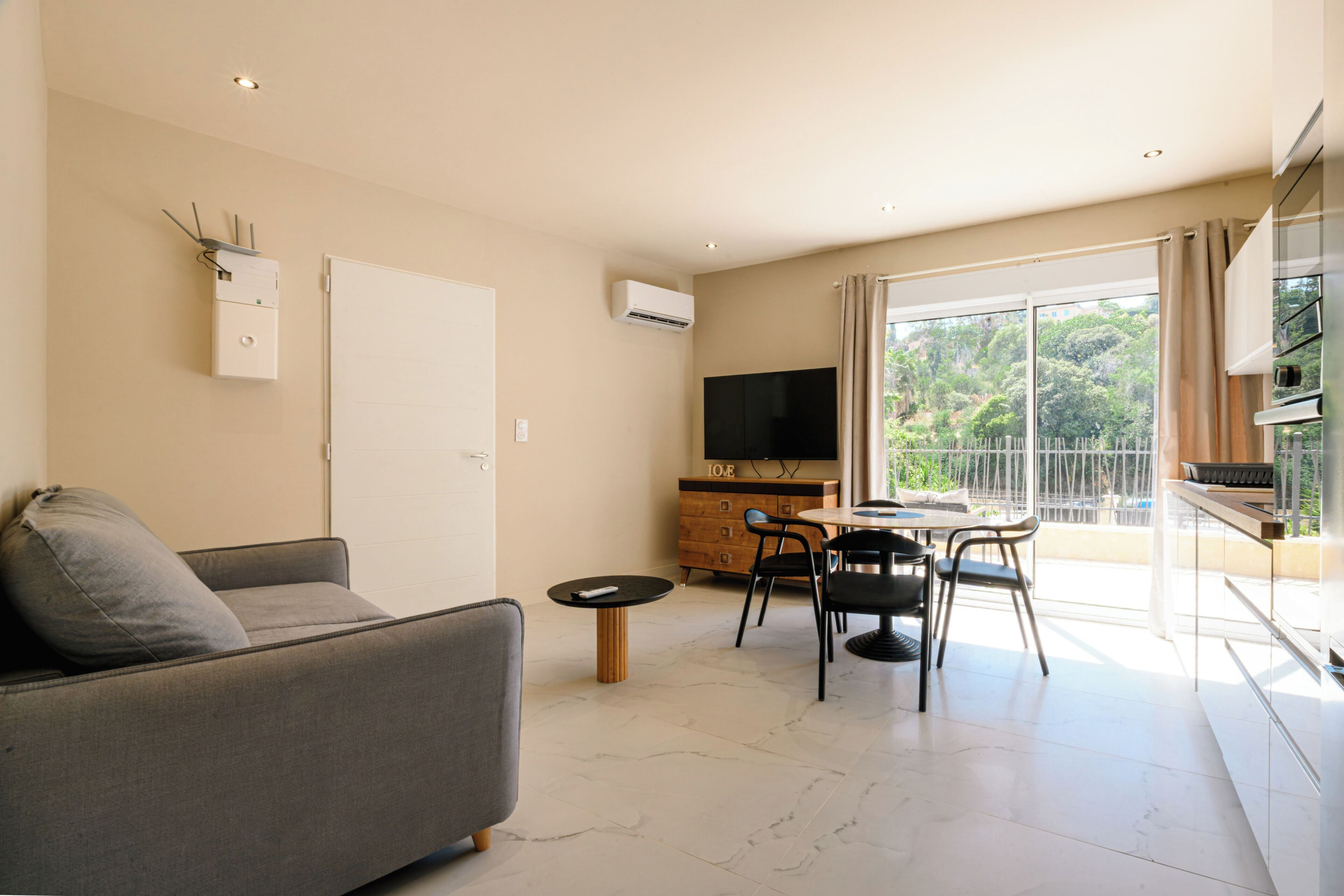 A modern living room with a sofa, a dining table with chairs, a flat-screen TV, and a sliding glass door leading to an outdoor balcony.