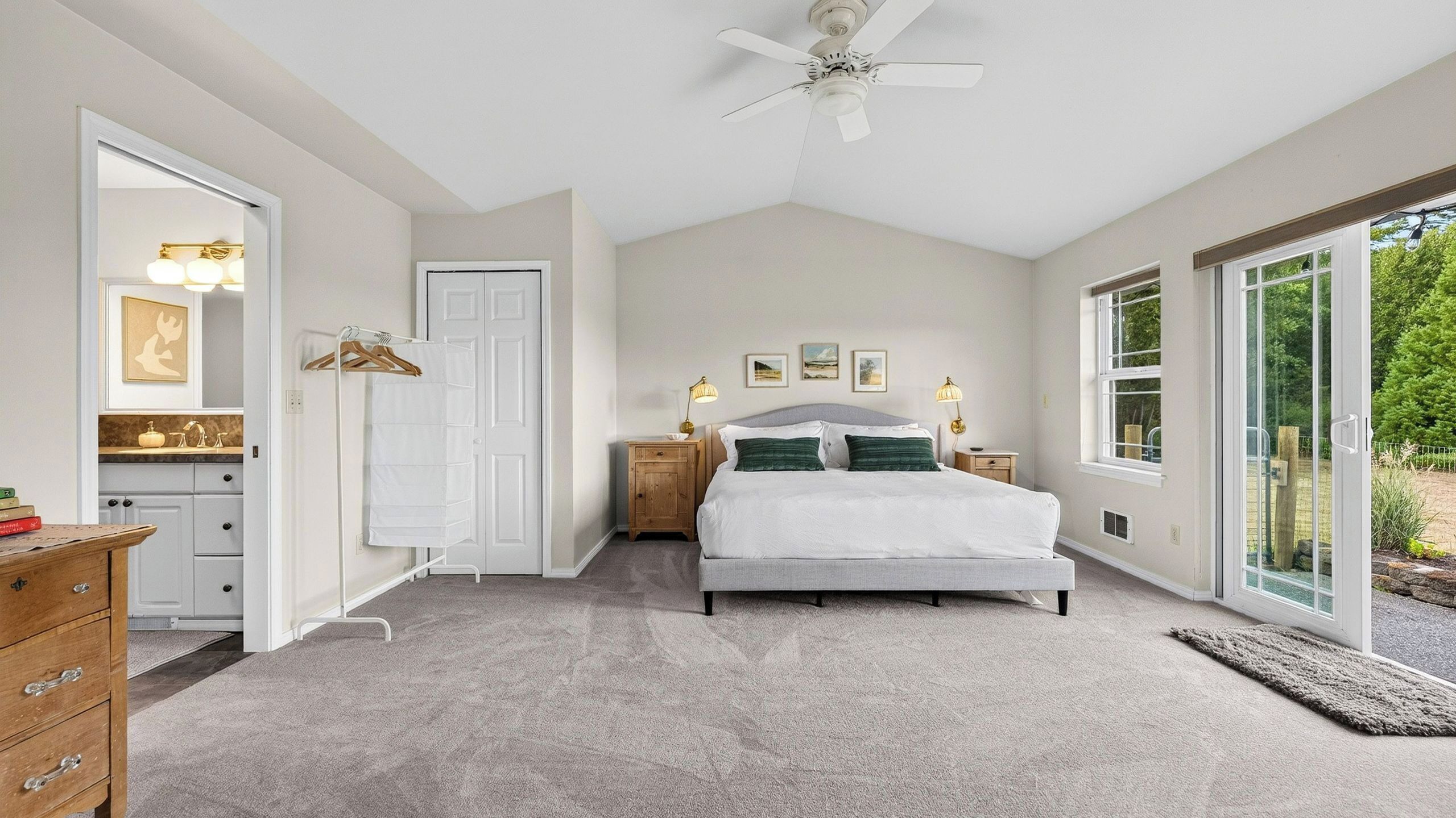 A bedroom with a bed, a dresser, a vanity, a ceiling fan, and a large window.