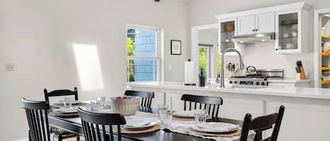 A dining area with a table set for four, a kitchen with white cabinets, and a window with a view of greenery.