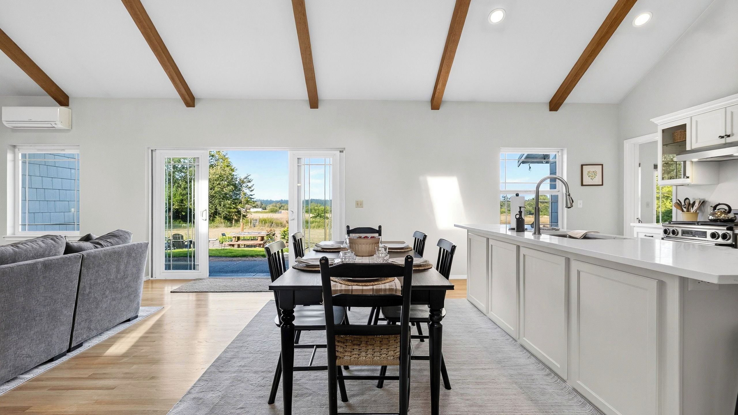 A modern kitchen with white cabinetry, a dining area with a table and chairs, and a view of the outdoors through large glass doors.