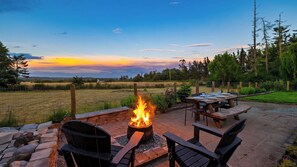 An outdoor patio with a fire pit, seating, and a view of a field and trees. - Edgefield Farmhouse by Avantstay Hot Tub + Firepit + 5 Private Acres Trailhead + Forts Nearby (Coupeville)