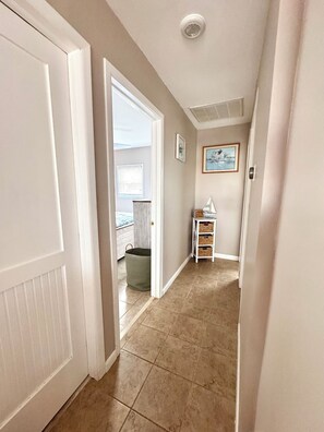 A hallway with a door on the left, a window with blinds, a small table with a lamp, and a trash can. - Mystic Island Lagoon Front Escape with Private Dock and Hot Tub ⚓️☀️ (Little Egg Harbor Township)