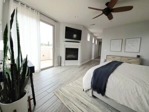 A bedroom with a bed, a ceiling fan, a fireplace, a TV, and a large window. - Luxury Coastal Retreat - Beach & Lagoon (Carlsbad)