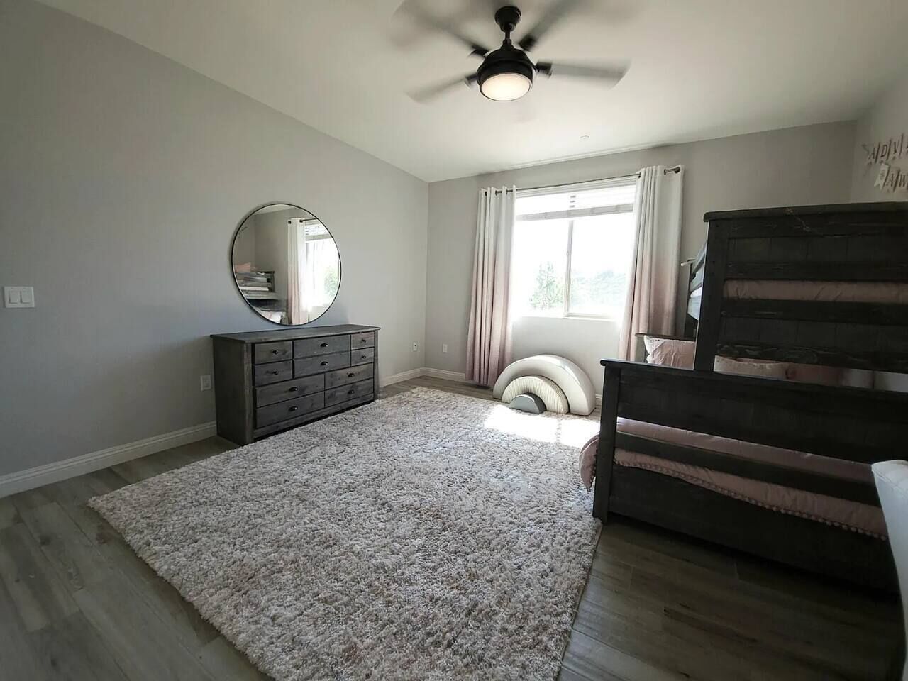 A bedroom with a ceiling fan, a dresser, a bed with a headboard, a window with curtains, and a rug.