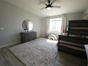 Une chambre avec un ventilateur de plafond, une commode, un lit avec une tête de lit, une fenêtre avec des rideaux et un tapis.