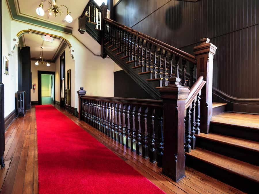 Un grand escalier recouvert d’un tapis rouge menant à un palier, flanqué de rampes en bois et surmonté d’un lustre.