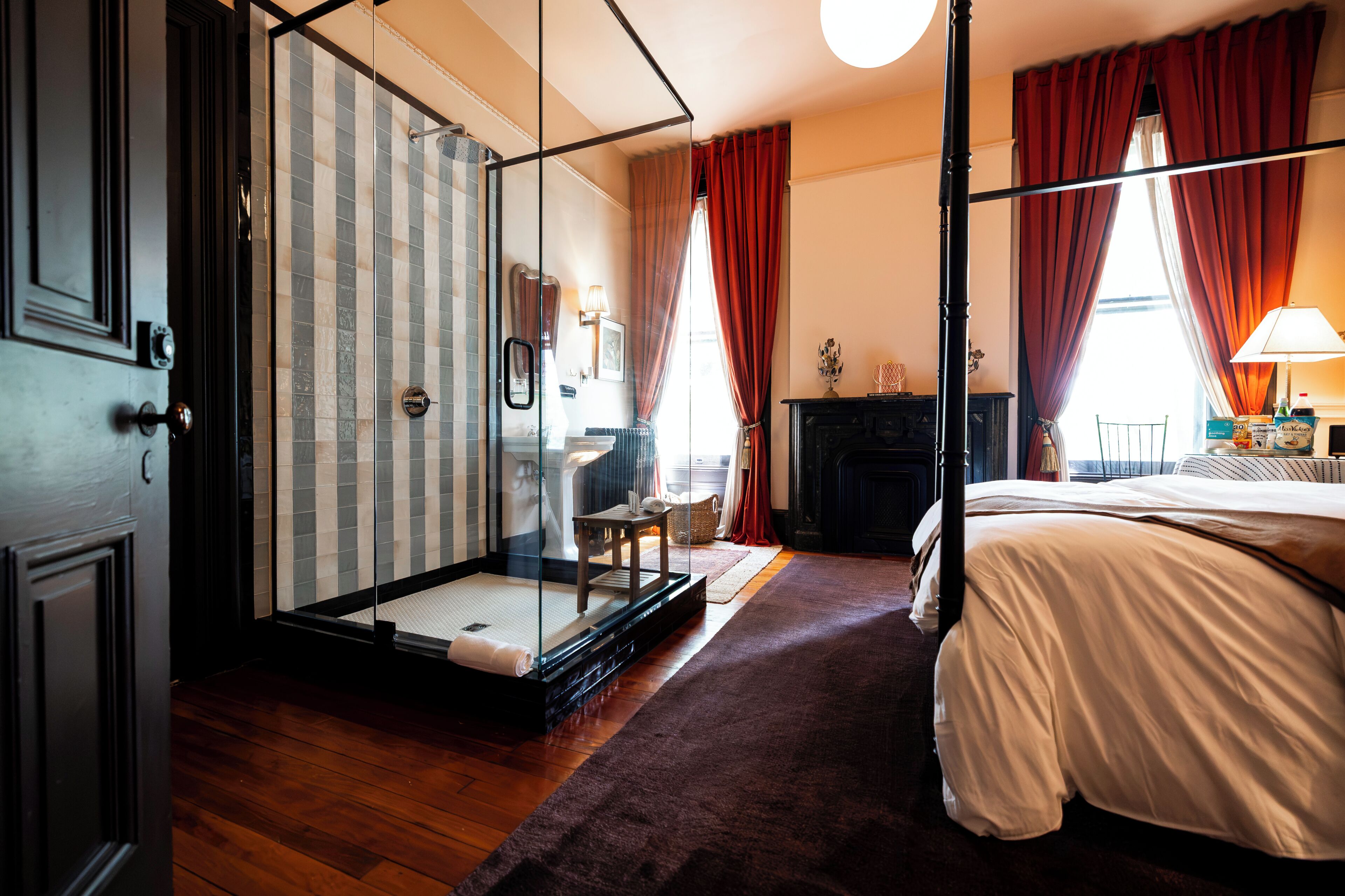 A hotel room with a glass-enclosed shower, a bed with white bedding, a fireplace, and a view of the city.
