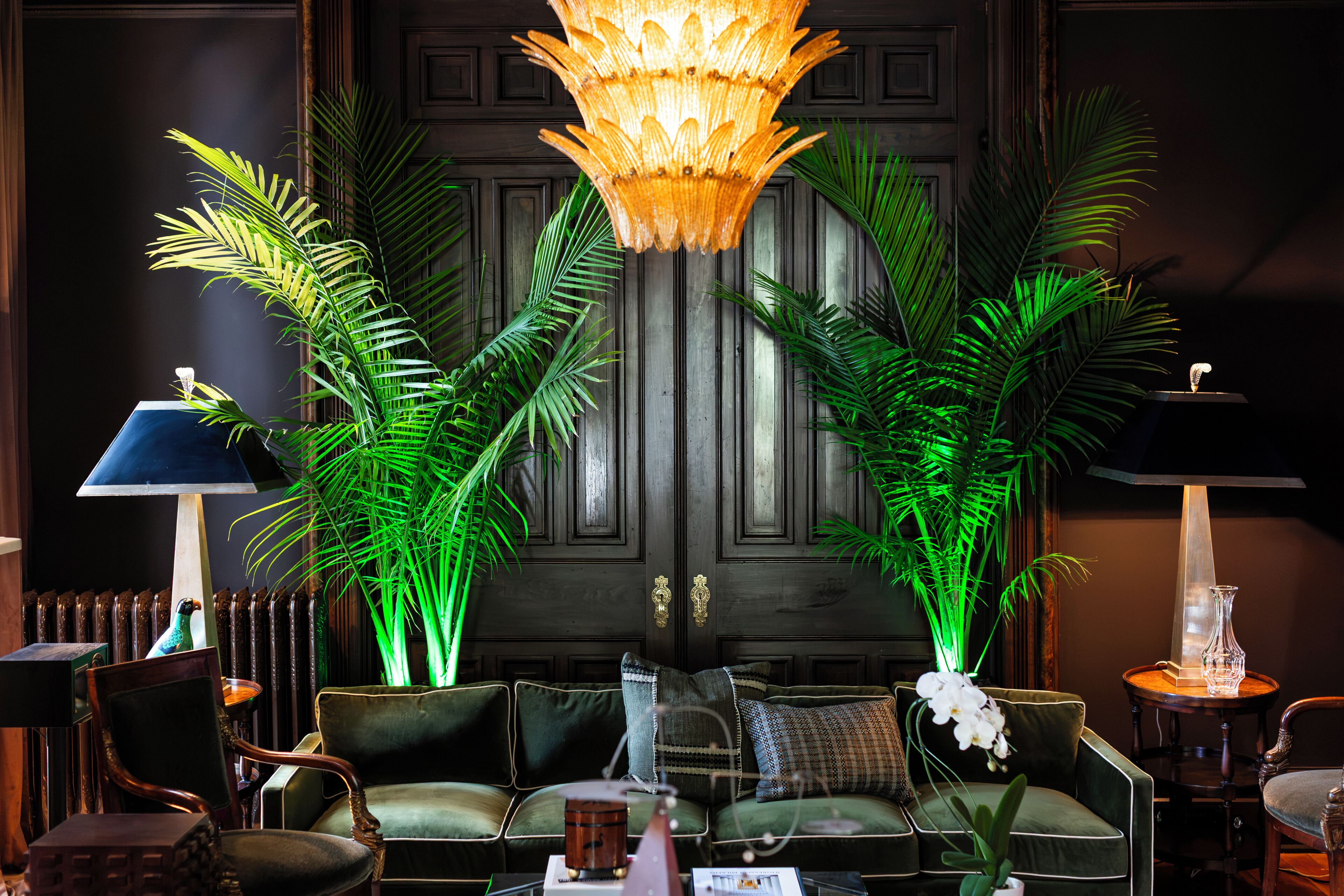 A room with a dark wooden door, a chandelier, green plants, and a seating area with a sofa and chairs.