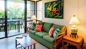 Un salon avec un canapé vert, une table basse en bois, des fauteuils en osier, une petite table avec une lampe et une grande fenêtre donnant sur un paysage verdoyant.