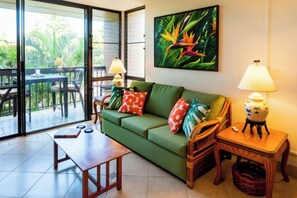 A living room with a green sofa, a wooden coffee table, wicker chairs, a small table with a lamp, and a large window with a view of greenery. - Tropical Maui Vista (Kihei)